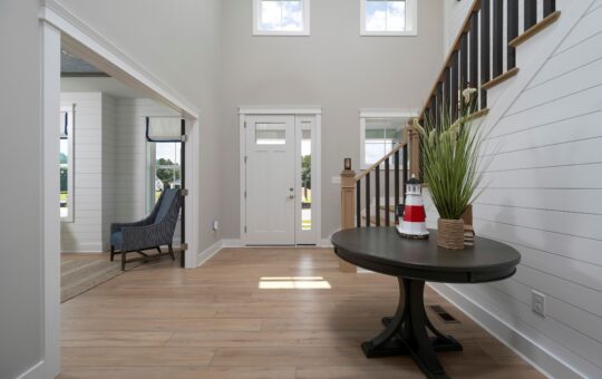 Bright entryway with light wood flooring, a round table holding a potted plant and a lighthouse figurine, white walls with shiplap, a staircase with dark railing, and double doors with windows above.