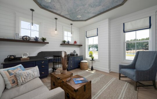 Bright home office with shiplap walls, large windows, blue cabinetry, a wooden desk, a beige sofa, a blue armchair, and a trunk-style coffee table; ceiling features a map design.