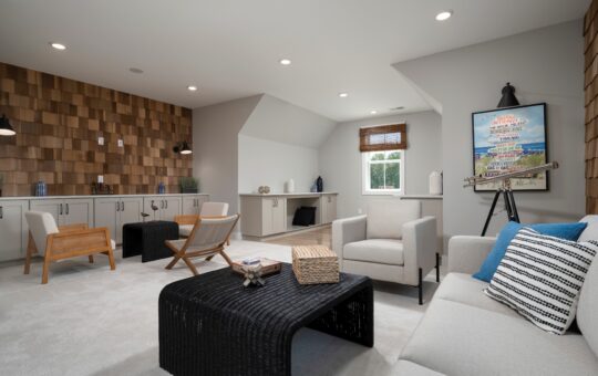 A modern, cozy living room with neutral tones features a sofa, armchairs, a black wicker coffee table, wall art, wood accent wall, built-in cabinets, and a window letting in natural light.