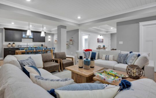 Modern open-concept living room and kitchen with neutral tones, cozy sofas, wooden coffee table with decor, games, and books, dining table with blue chairs, and bright overhead lighting.