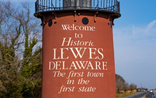 A red lighthouse-shaped sign reads “Welcome to Historic Lewes Delaware, The first town in the first state,” standing by a road with trees and blue sky in the background.