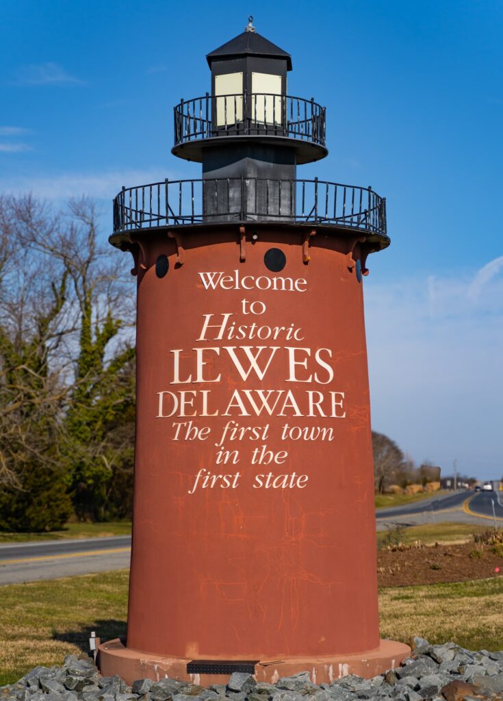 A red lighthouse-shaped sign reads “Welcome to Historic Lewes Delaware, The first town in the first state,” standing by a road with trees and blue sky in the background.