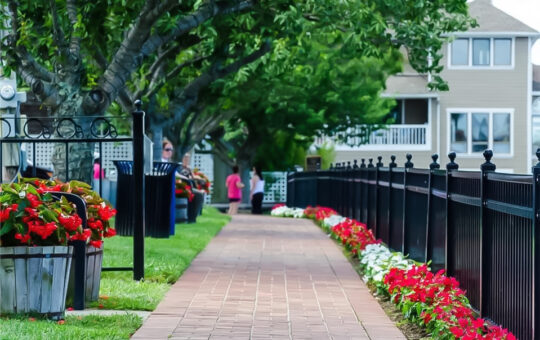 A brick walkway lined with red and white flowers runs parallel to a black metal fence, with trees and houses in the background and a few people walking.
