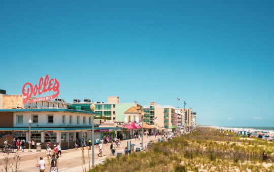 A sunny beach boardwalk scene with people walking, sandy dunes, ocean waves, and colorful buildings, including a shop with a large red Dolle’s Salt Water Taffy sign on the roof.