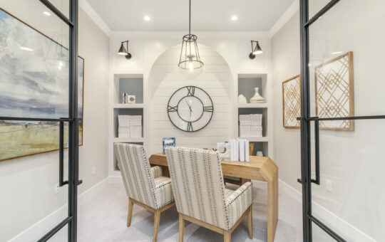 A modern home office with a wooden desk, two patterned chairs, built-in shelves, wall art, and a large decorative clock on a shiplap accent wall. Glass doors and soft lighting set the scene for implementing tips for organizing your home office.