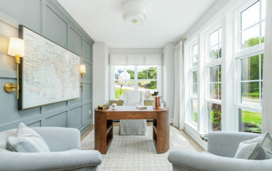 Bright, modern home office with large windows, a wooden desk, and two light gray armchairs. Sunlight fills the space, highlighting the clean design—an inspiring setting for anyone seeking tips for organizing your home office.