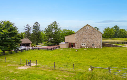 A large stone barn with a tan upper section stands in a green fenced pasture with sheep grazing. Trees and a small open structure are visible behind the barn under a clear blue sky.