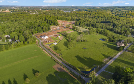 Aerial view of a rural landscape with green fields, scattered houses, a winding road, and an area under construction surrounded by trees and forests under a blue sky with few clouds.