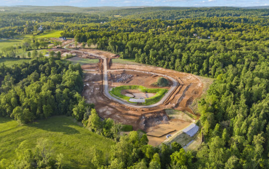 Aerial view of a construction site surrounded by dense green forest, with cleared earth forming winding paths and a large central area, under a bright sky with scattered clouds.