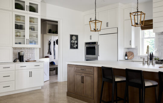 Modern kitchen with white cabinets, a built-in oven, wooden island with black chairs, pendant lights, and a pantry in the background. Neutral tones and natural light create a clean, inviting atmosphere.