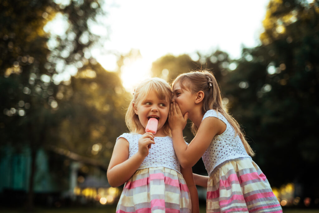 Shot of a two young girls whispering while eating ice cream outdoors at sunset.