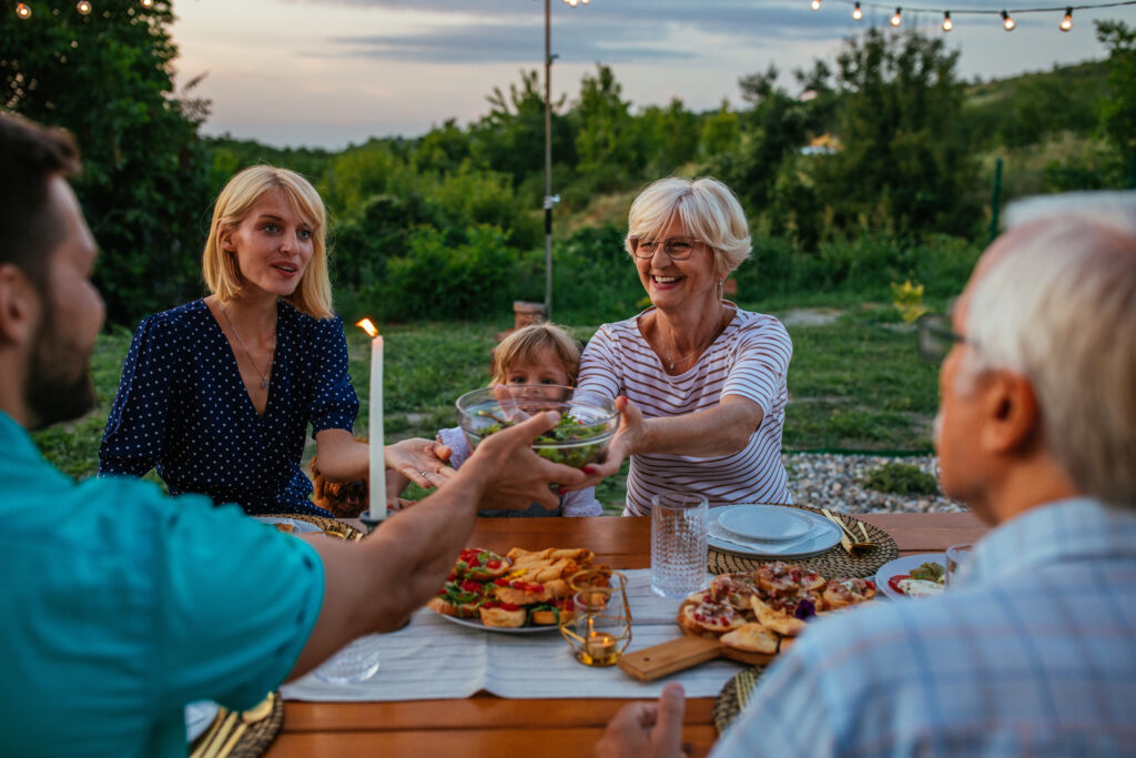 Three generations family sitting at the dining table in the backyard. Little girl enjoying dinner with her parents and grandparents outdoors.