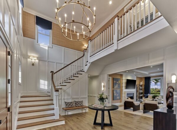 A bright, two-story foyer with a large round chandelier, wooden staircase with white railings, light wood floors, and an open view into a cozy living room with a fireplace.