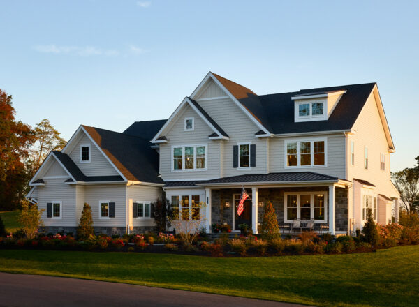 A large, modern two-story house with gray siding, stone accents, a front porch, and well-manicured landscaping. An American flag hangs by the entrance, and the home is lit by warm evening sunlight.