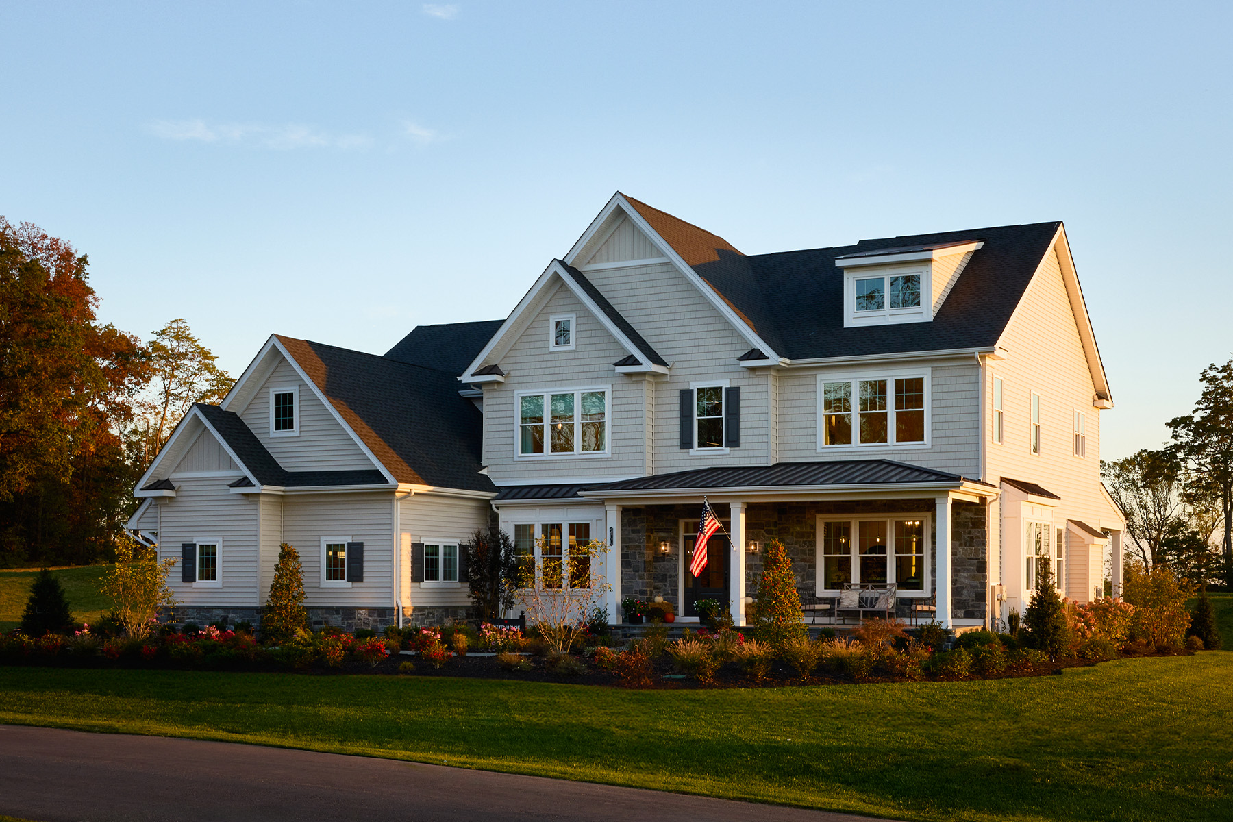A large, modern two-story house with gray siding, stone accents, a front porch, and well-manicured landscaping. An American flag hangs by the entrance, and the home is lit by warm evening sunlight.