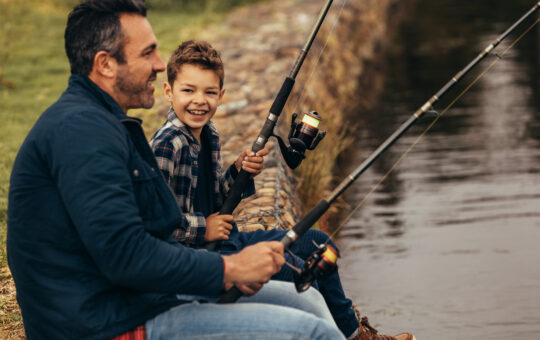 A man and a smiling boy sit side by side on a stone edge, holding fishing rods and fishing in a calm body of water, enjoying a relaxed outdoor moment together.