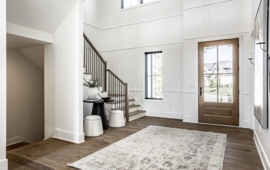 A bright, modern entryway with a wooden front door, tall windows, light walls, a staircase with a wooden handrail, a textured area rug, and a small table with two white stools beneath it.