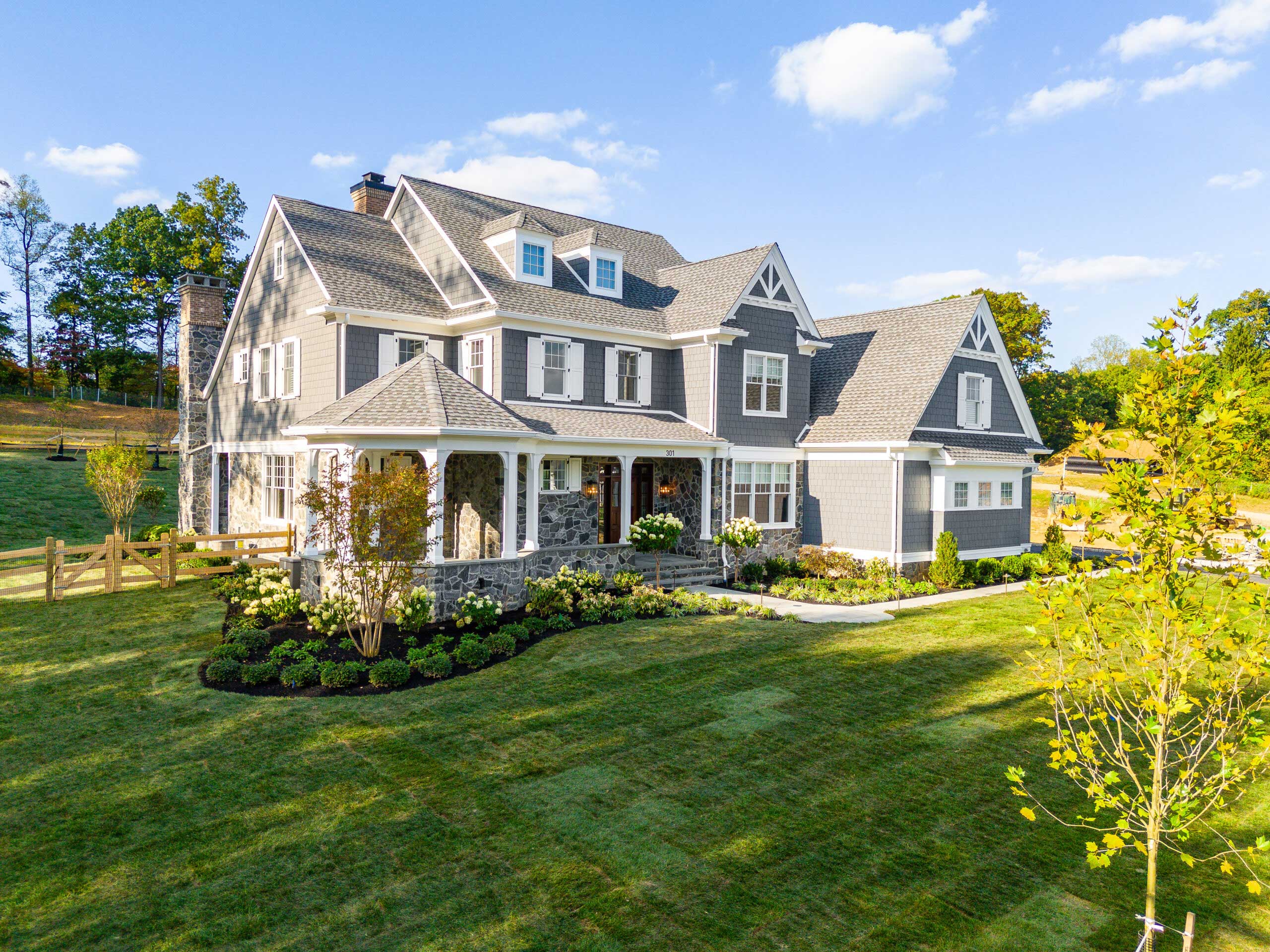A large, two-story gray house with white trim and a wraparound porch sits on a well-manicured lawn with shrubs, trees, and a wooden fence, under a blue sky with scattered clouds.