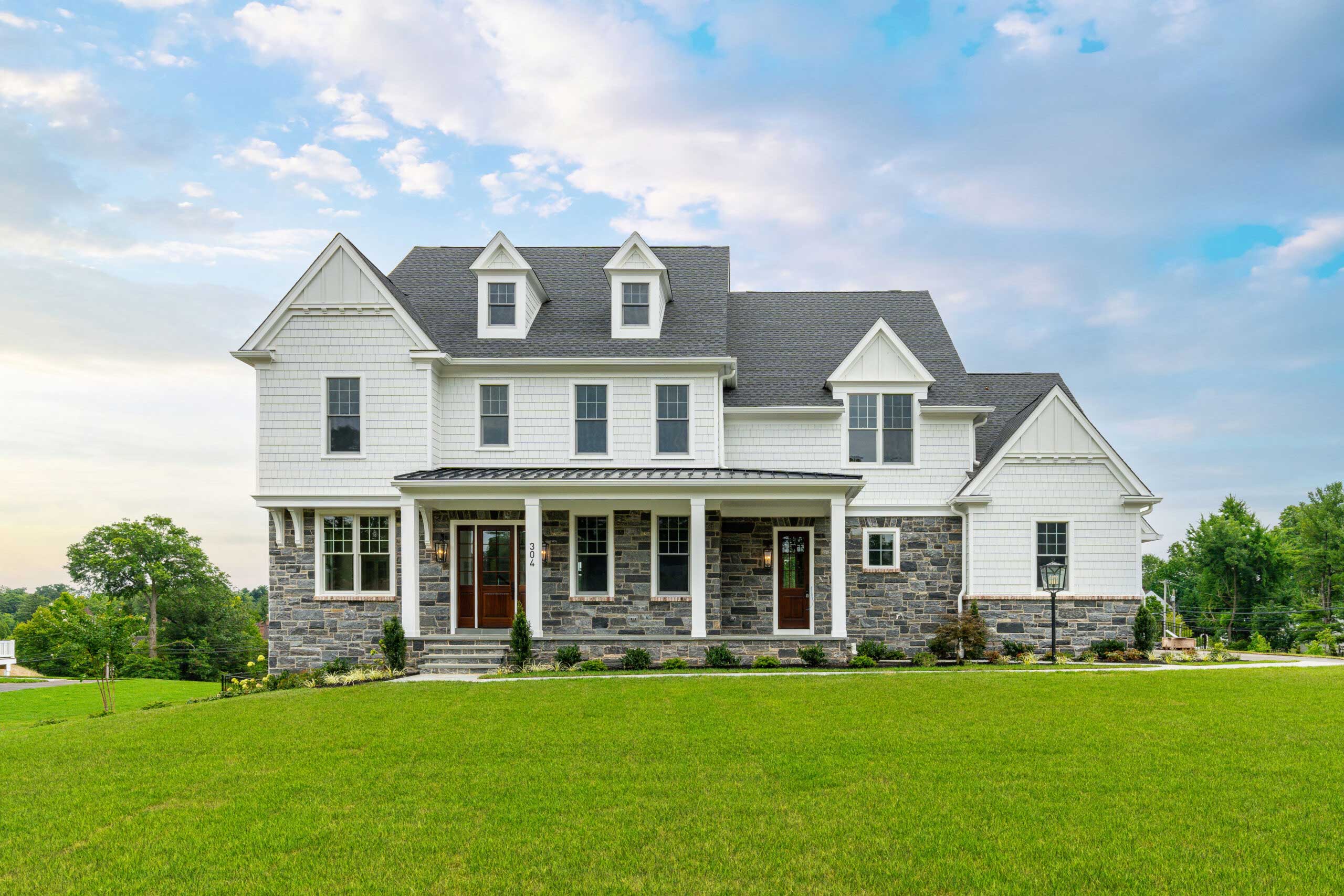 A large, modern two-story house with white siding, stone accents, multiple gables, and a wide front porch, surrounded by a well-manicured green lawn under a partly cloudy sky.