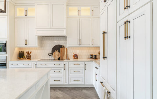 Modern kitchen with white cabinets, gold handles, white countertops, and a tile backsplash. Wooden cutting boards, utensils, and jars add decor. Natural light brightens the clean and organized space.