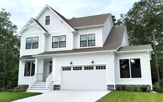 A modern two-story white house with black trim, a front porch, double garage, and large windows, sits on a driveway surrounded by greenery and trees. The sky is partly cloudy.