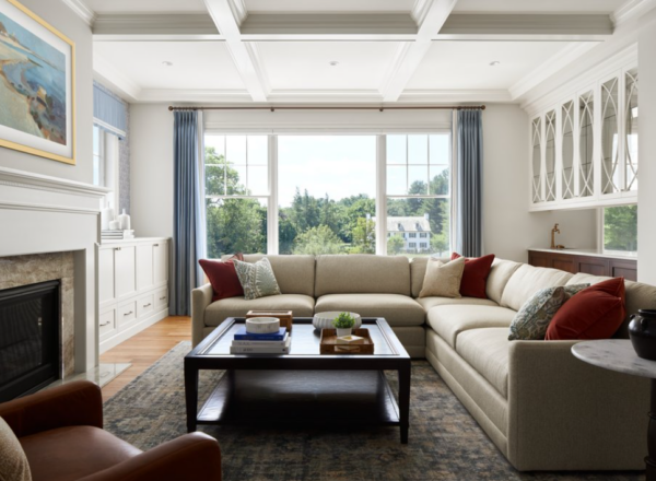 A bright living room with a large beige sectional sofa, red and gray pillows, a dark coffee table, a fireplace, and a big window with blue curtains overlooking a green, tree-filled view.
