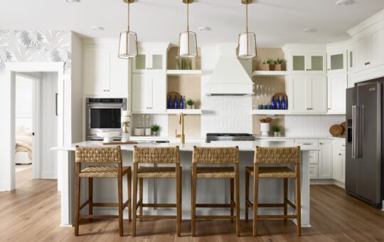 Modern kitchen with white cabinets, three pendant lights, a large island with four woven bar stools, stainless steel appliances, wooden shelves, and decorative blue glass bottles. Light wood flooring completes the space.