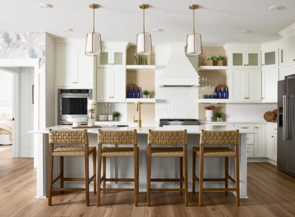 Modern kitchen with white cabinets, three pendant lights, a large island with four woven bar stools, stainless steel appliances, wooden shelves, and decorative blue glass bottles. Light wood flooring completes the space.