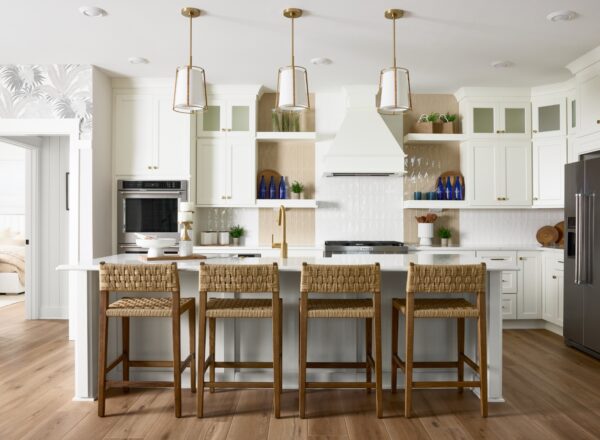 Modern kitchen with white cabinets, three pendant lights, a large island with four woven bar stools, stainless steel appliances, wooden shelves, and decorative blue glass bottles. Light wood flooring completes the space.