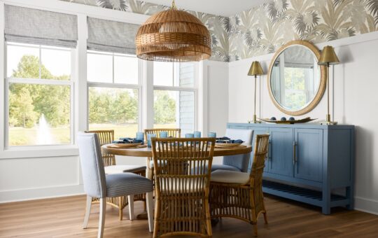 Bright dining room with large windows, a round wooden table surrounded by wicker and upholstered chairs, a woven pendant lamp, blue sideboard, round mirror, and patterned wallpaper. Trees and water fountain visible outside.