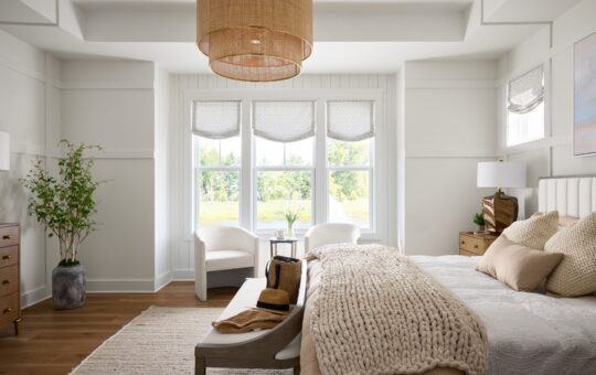 A bright, airy bedroom with large windows, neutral decor, a cozy bed with a chunky knit blanket, two armchairs by the window, wooden furniture, and a woven pendant light. Greenery in a vase adds a natural touch.