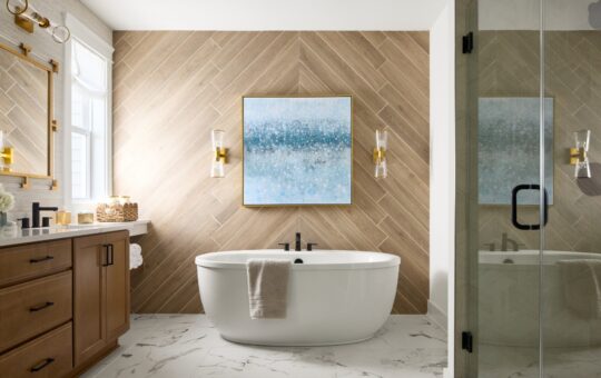 Modern bathroom with a white freestanding tub, wood-patterned accent wall, large blue abstract art, marble floor, wooden vanity with double sinks, gold fixtures, glass shower, and a window letting in natural light.