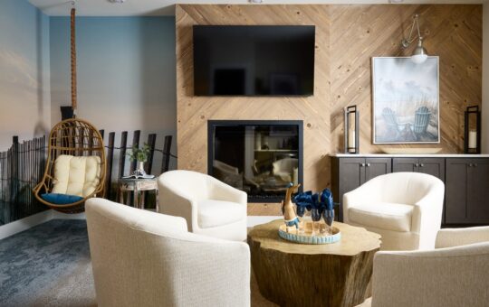 A modern living room with four white armchairs around a wooden coffee table, a fireplace below a wall-mounted TV, a beach scene mural, a hanging wicker chair, and coastal décor accents.