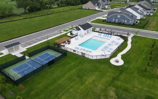 Aerial view of a residential community with a fenced tennis court, swimming pool area with lounge chairs, small building, parking lot, and surrounding green lawns. Nearby houses are visible along paved streets.