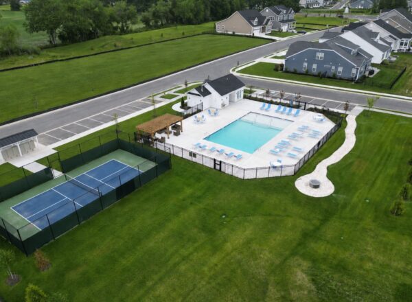Aerial view of a residential community with a fenced tennis court, swimming pool area with lounge chairs, small building, parking lot, and surrounding green lawns. Nearby houses are visible along paved streets.