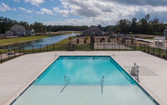 A large outdoor swimming pool with clear blue water is surrounded by a concrete deck and fencing. In the background, there are houses, trees, and a pond under a partly cloudy sky.