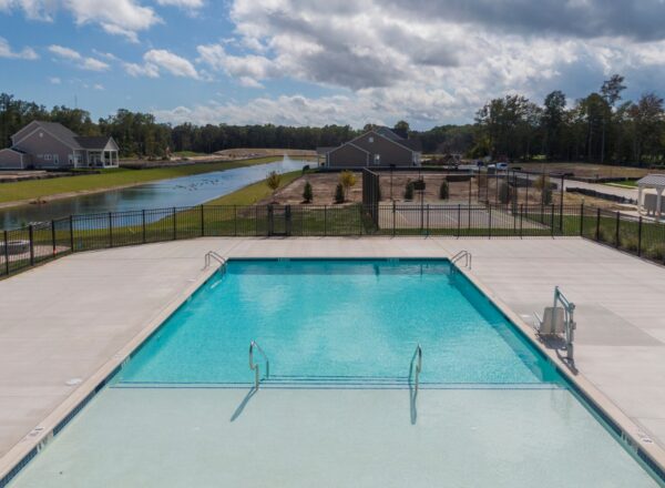 A large outdoor swimming pool with clear blue water is surrounded by a concrete deck and fencing. In the background, there are houses, trees, and a pond under a partly cloudy sky.