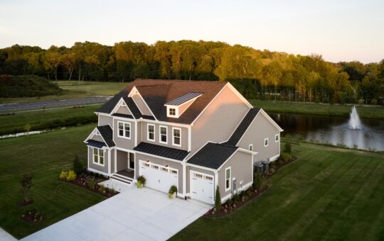 A large two-story suburban house with gray siding and white trim sits next to a pond with a fountain, surrounded by a green lawn and trees under a clear sky at sunset.