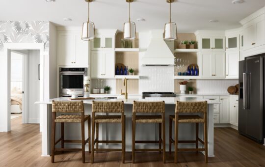 Modern white kitchen with a large island, four woven barstools, brass pendant lights, stainless steel appliances, open shelves with blue glass bottles, and wood accents on light hardwood floors.
