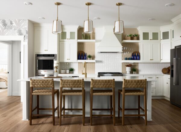 Modern white kitchen with a large island, four woven barstools, brass pendant lights, stainless steel appliances, open shelves with blue glass bottles, and wood accents on light hardwood floors.