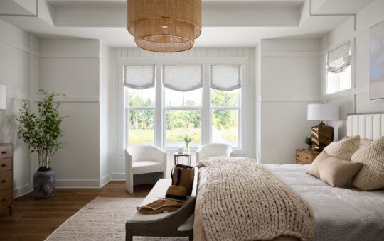 Bright, modern bedroom with large windows, neutral decor, a knitted blanket on the bed, two armchairs, light wood furniture, a potted plant, and a woven pendant light. Sunlight streams in, creating a cozy, inviting feel.