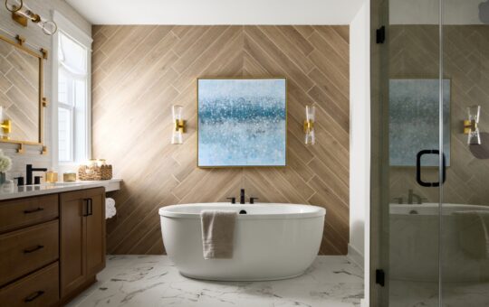 A modern bathroom featuring a freestanding white bathtub with a towel draped over the edge, a wooden vanity, a large blue abstract painting, and marble floor tiles. Light streams in from a window on the left.