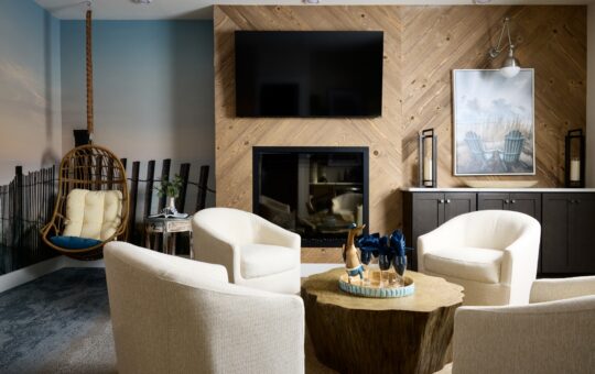 A cozy living room with four cream armchairs around a wooden coffee table, a fireplace with a TV above, a hanging wicker chair, and coastal-themed wall art and decor.