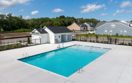 A rectangular outdoor swimming pool with clear blue water is surrounded by a white concrete deck, a small white pool house, and nearby residential houses under a blue sky with scattered clouds.