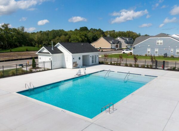 A rectangular outdoor swimming pool with clear blue water is surrounded by a white concrete deck, a small white pool house, and nearby residential houses under a blue sky with scattered clouds.