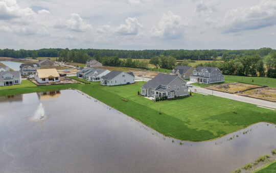 Aerial view of a suburban neighborhood with newly built houses near a pond with a fountain, green lawns, and nearby construction, surrounded by trees and open fields under a partly cloudy sky.