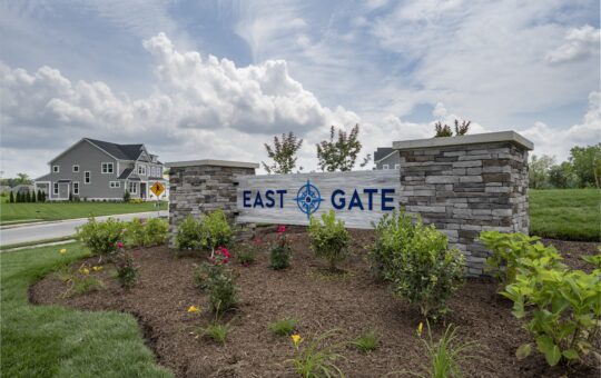 A stone sign reading East Gate stands in front of landscaped flowers, with suburban houses and a cloudy sky in the background.