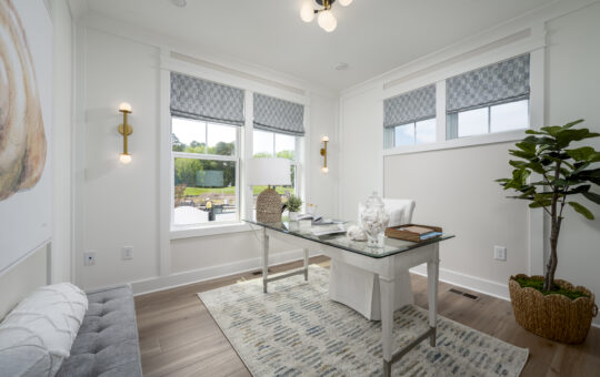 A bright, modern home office with a glass desk, white chair, potted plant, patterned rug, and large windows letting in natural light. Decorative lights and artwork accent the clean, airy space.