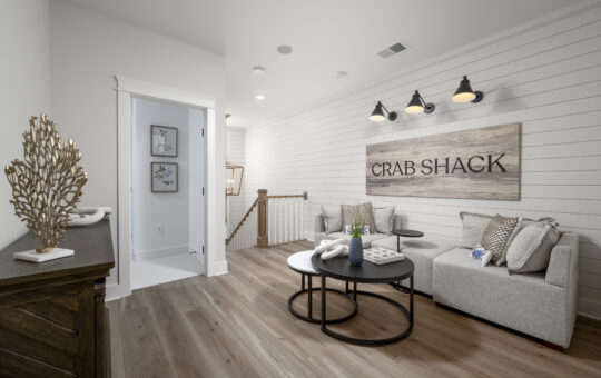 A cozy, coastal-themed sitting area with a light gray sectional sofa, round black coffee tables, and a “Crab Shack” sign on a shiplap wall. Nautical decor and soft wood floors complete the airy space.