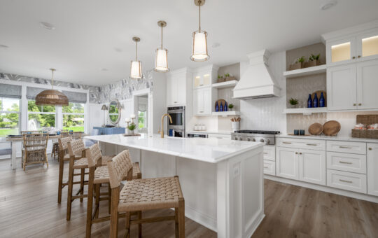 Bright, modern kitchen with white cabinets, island with four woven stools, pendant lights, open shelves, stainless steel appliances, wooden flooring, and a dining area with wicker chairs by large windows.